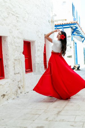 Téléchargez les photos : Humeur de vacances. femme brune en robe d'été rouge se tient à la mode près de fond de mur blanc avec des villas grecques avec portes bleues et citronnier à la journée ensoleillée. concept de voyage, espace libre - en image libre de droit