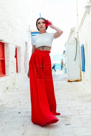 Téléchargez les photos : Charme méditerranéen : Une femme brune en robe rouge se dresse gracieusement contre un mur blanc avec des villas grecques, capturant l'allure d'une journée ensoleillée - en image libre de droit