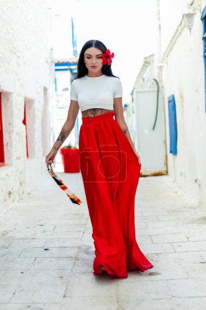Téléchargez les photos : Aventure grecque : Une femme brune élégante dans une robe rouge se tient à la mode contre un mur blanc avec des villas grecques, rayonnant des vibrations de vacances - en image libre de droit