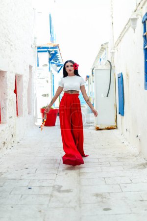 Téléchargez les photos : Ambiances de voyage : Une brune en robe d'été rouge pose sur un fond de mur blanc avec des villas grecques, embrassant l'ambiance de vacances avec flair - en image libre de droit