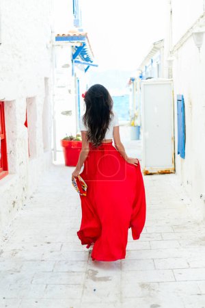 Téléchargez les photos : Fuite en Grèce : Une brune élégante dans une robe rouge pose sur un fond de mur blanc avec des villas grecques, incarnant l'essence d'une escapade ensoleillée - en image libre de droit