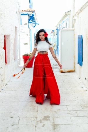 Téléchargez les photos : Escapade grecque : Une brune élégante dans une robe rouge se dresse contre un mur blanc avec des villas grecques, incarnant l'essence des jours de vacances ensoleillés - en image libre de droit