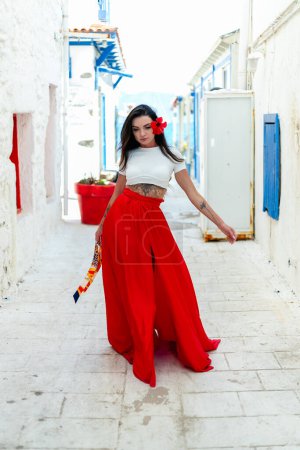 Téléchargez les photos : Ambiances de voyage : Une brune en robe d'été rouge pose sur un fond de mur blanc avec des villas grecques, embrassant l'ambiance de vacances avec flair - en image libre de droit