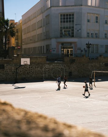 Foto de Cádiz, Andalucía, España; 25 de noviembre de 2023: Siluetas de adolescentes jugando al baloncesto en un día soleado. Amigos jugando baloncesto en una cancha urbana. - Imagen libre de derechos