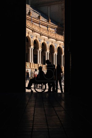 Foto de Sevilla, España; 25 de enero de 2024: Silueta de un hombre empujando una silla de ruedas. Silueta de un hombre en silla de ruedas con la Plaza de España al fondo. - Imagen libre de derechos