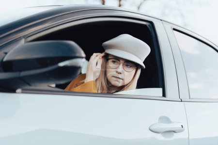 Téléchargez les photos : Une femme d'âge moyen portant des lunettes et un chapeau est assise dans une voiture et regarde son visage dans le rétroviseur - en image libre de droit