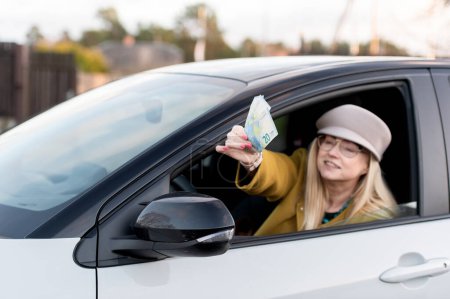 Photo for Smiling middle-aged woman gives euro banknotes through the window of a modern luxury car - Royalty Free Image