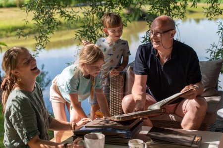 Téléchargez les photos : Grand-père avec ses petits-enfants regardant de vieux albums de photos sur la terrasse extérieure par une journée d'été ensoleillée - en image libre de droit