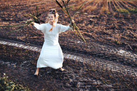 Téléchargez les photos : Belle femme en robe danse pieds nus sur un champ boueux, tenant des roseaux dans ses mains - en image libre de droit
