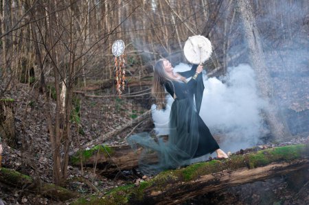 Téléchargez les photos : Femme sauvage jouant du tambour chamanique debout sur le tronc vert d'arbre de mousse dans la forêt, bombe blanche de fumée - en image libre de droit