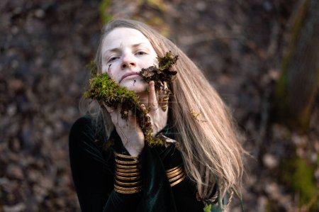 Female shaman performs a nature ritual by smearing her face with forest moss and soil