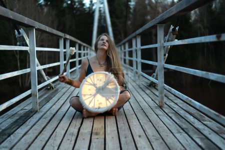 Shaman female playing  shamanic drum on wooden bridge, golden bracelet and rings