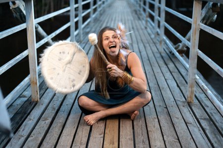 Divertido chamán femenino tocando tambor chamánico en puente de madera, expresión de la cara