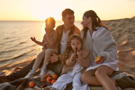 Foto de Una familia joven con dos niños sentados en una manta en un picnic en una orilla arenosa junto al mar al atardecer divirtiéndose y relajándose el fin de semana. - Imagen libre de derechos