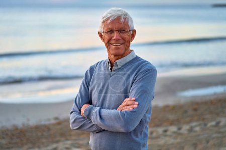 Photo for An elderly intelligent man in glasses walking along the beach near the sea in autumn in cool weather. - Royalty Free Image