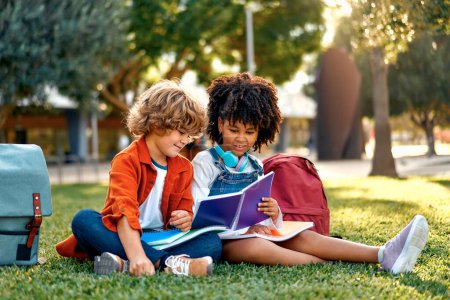 Téléchargez les photos : Adorables écoliers faisant leurs devoirs ensemble tout en étant assis sur l'herbe dans le parc. Je retourne à l'école. afro-américaine fille avec caucasien mignon garçon communiquer après l'école. - en image libre de droit