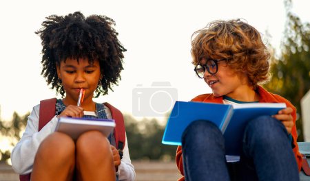 Charming smart schoolchildren doing their homework together while sitting on the stairs near the school. Back to school. African American girl with caucasian boy in glasses studying together.