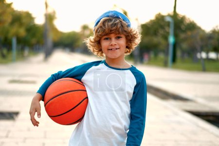 Sports and basketball. Stylish cute boy standing with a basketball ball outdoors in the park. Concept of sport, movement, healthy lifestyle, ad, action, motion.