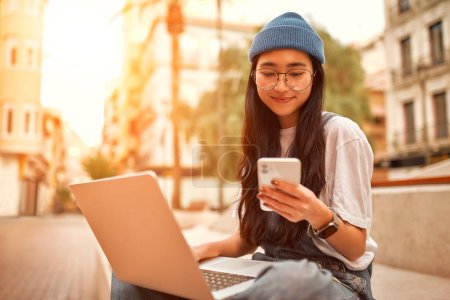 Porträt einer asiatischen Touristin, die im weißen Hemd auf einer Bank sitzt und mit einem Laptop telefoniert. Studentinnen studieren in der Nähe des Campus. Freiberufler arbeiten auf der Straße.
