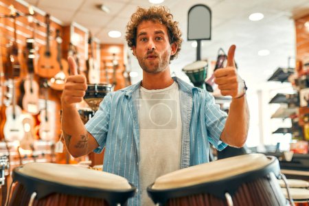 Foto de Guapo barbudo rizado tocando la batería con las manos en una tienda de instrumentos musicales antes de comprar y mostrar un gesto de pulgar hacia arriba. - Imagen libre de derechos