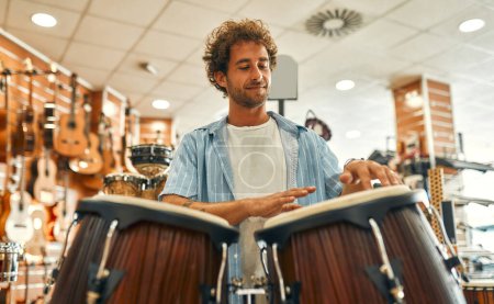 Foto de Guapo barbudo rizado tocando la batería con las manos en una tienda de instrumentos musicales antes de comprar. - Imagen libre de derechos
