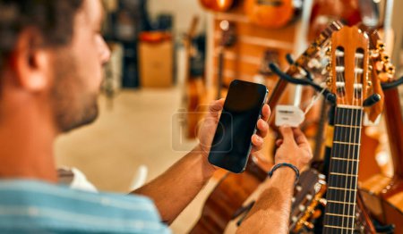 Foto de Handsome bearded man scanning a code on a price tag using his smartphone in a musical instrument store. - Imagen libre de derechos