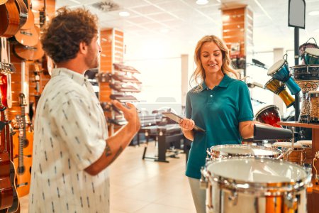 Foto de Handsome bearded curly man choosing a drum in a musical instrument store and consulting a consultant before buying. - Imagen libre de derechos