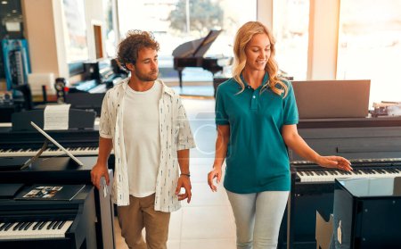 Foto de Handsome bearded curly-haired man choosing a piano in a musical instrument store getting the help of a consultant. Woman seller telling the buyer about the piano. - Imagen libre de derechos