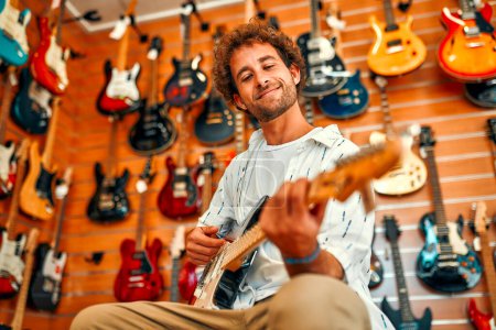 Foto de Handsome bearded curly man in front of walls with many electric guitars in a musical instrument store and trying to play it. Hobbies and recreation. Buying a guitar in a store. - Imagen libre de derechos