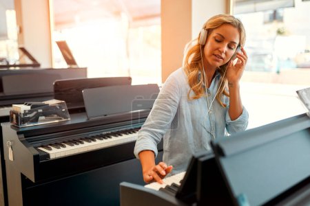Foto de Joven mujer hermosa con auriculares sentados al piano y tocando en una tienda de instrumentos musicales. Pasatiempos y recreación. Comprar un piano en una tienda. - Imagen libre de derechos