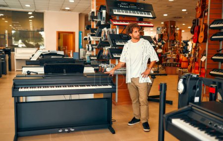 Foto de Young attractive man choosing a piano or synthesizer in a musical instrument store. Hobbies and recreation. Buying a midi keyboard in a store. - Imagen libre de derechos