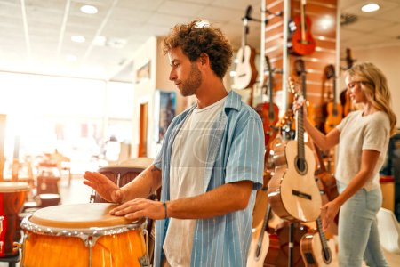 Foto de Un hombre tocando la batería con las manos en una tienda de instrumentos musicales. Mujer en el fondo eligiendo una guitarra en una tienda de música. - Imagen libre de derechos