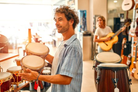 Foto de A man playing drums with his hands in a musical instrument store. Woman in the background choosing a guitar in a music store. - Imagen libre de derechos