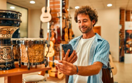 Foto de Hombre barbudo guapo escaneando un código en una etiqueta de precio usando su teléfono inteligente en una tienda de instrumentos musicales. - Imagen libre de derechos