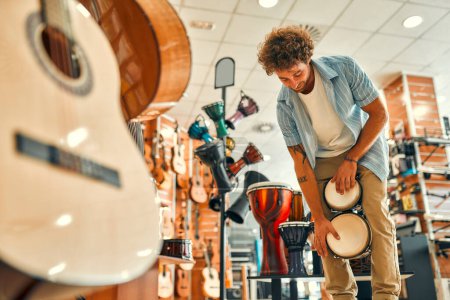 Foto de Guapo barbudo rizado tocando tambores pequeños con las manos en una tienda de instrumentos musicales antes de comprar. - Imagen libre de derechos