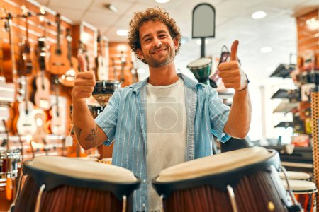 Foto de Guapo barbudo rizado tocando la batería con las manos en una tienda de instrumentos musicales antes de comprar y mostrar un gesto de pulgar hacia arriba. - Imagen libre de derechos