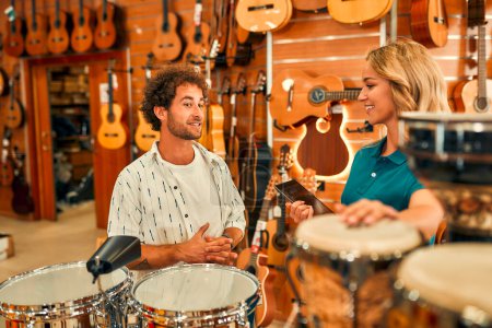 Foto de Guapo barbudo rizado hombre elegir un tambor en una tienda de instrumentos musicales y consultar a un consultor antes de comprar. - Imagen libre de derechos