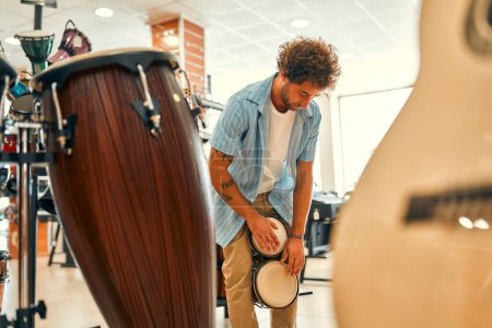 Foto de Handsome bearded curly man playing small drums with his hands in a musical instrument store before buying. - Imagen libre de derechos