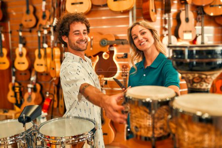 Foto de Handsome bearded curly man choosing a drum in a musical instrument store and consulting a consultant before buying. - Imagen libre de derechos
