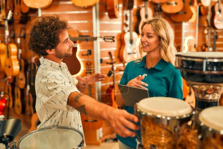 Foto de Guapo barbudo rizado hombre elegir un tambor en una tienda de instrumentos musicales y consultar a un consultor antes de comprar. - Imagen libre de derechos