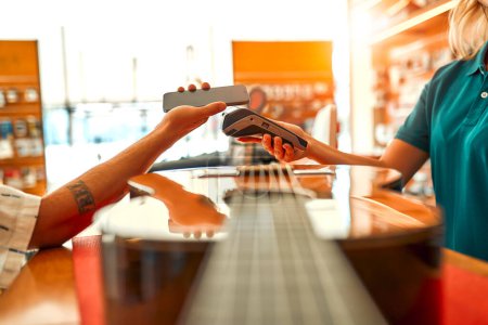 Foto de Handsome bearded curly man buying a guitar in a musical instrument store and paying for the purchase using a smartphone. Contactless payment. Woman seller selling a guitar to a buyer. - Imagen libre de derechos