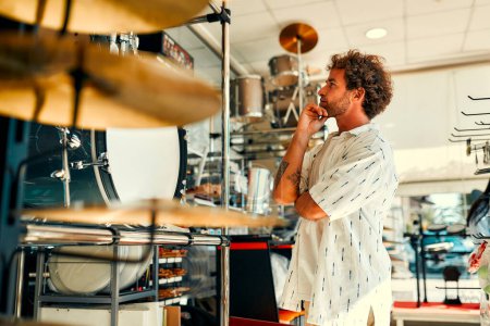 Foto de Guapo barbudo rizado hombre elegir un tambor en una tienda de instrumentos musicales. Pasatiempos y recreación. Un hombre comprando un tambor en una tienda. - Imagen libre de derechos