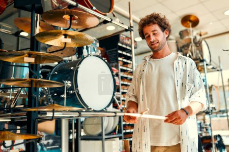 Foto de Handsome bearded curly man choosing a drum in a musical instrument store. Hobbies and recreation. A man buying a drum in a store and trying to play it. - Imagen libre de derechos