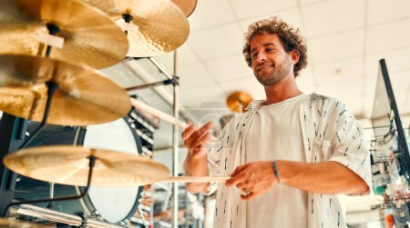 Foto de Handsome bearded curly man choosing a drum kit in a musical instrument store. Hobbies and recreation. A man buying a drum in a store and trying to play it. - Imagen libre de derechos