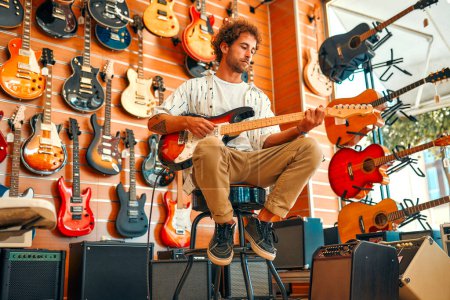 Foto de Guapo barbudo rizado hombre en frente de las paredes con muchas guitarras eléctricas en una tienda de instrumentos musicales y tratando de tocarlo. Pasatiempos y recreación. Comprar una guitarra en una tienda. - Imagen libre de derechos