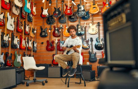 Foto de Guapo barbudo rizado hombre en frente de las paredes con muchas guitarras eléctricas en una tienda de instrumentos musicales y tratando de tocarlo. Pasatiempos y recreación. Comprar una guitarra en una tienda. - Imagen libre de derechos