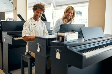 Foto de Young couple sitting at the piano and playing four hands in a musical instrument store. Hobbies and recreation. Buying a piano in a store. - Imagen libre de derechos