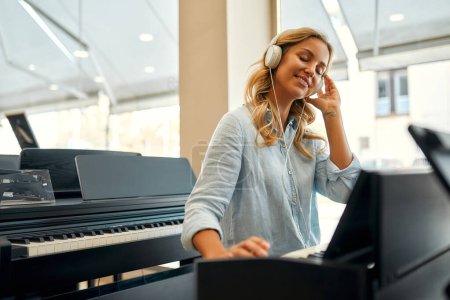 Foto de Young beautiful woman in headphones sitting at the piano and playing it in a musical instrument store. Hobbies and recreation. Buying a piano in a store. - Imagen libre de derechos