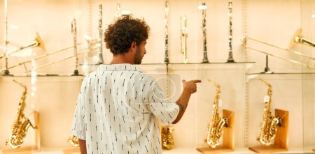 Foto de A young attractive man standing in front of a showcase with wind instruments in a musical instrument store. Hobbies and recreation. - Imagen libre de derechos