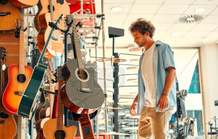 Foto de Guapo barbudo rizado hombre elegir una guitarra en una tienda de instrumentos musicales. Aficiones y recreación. - Imagen libre de derechos